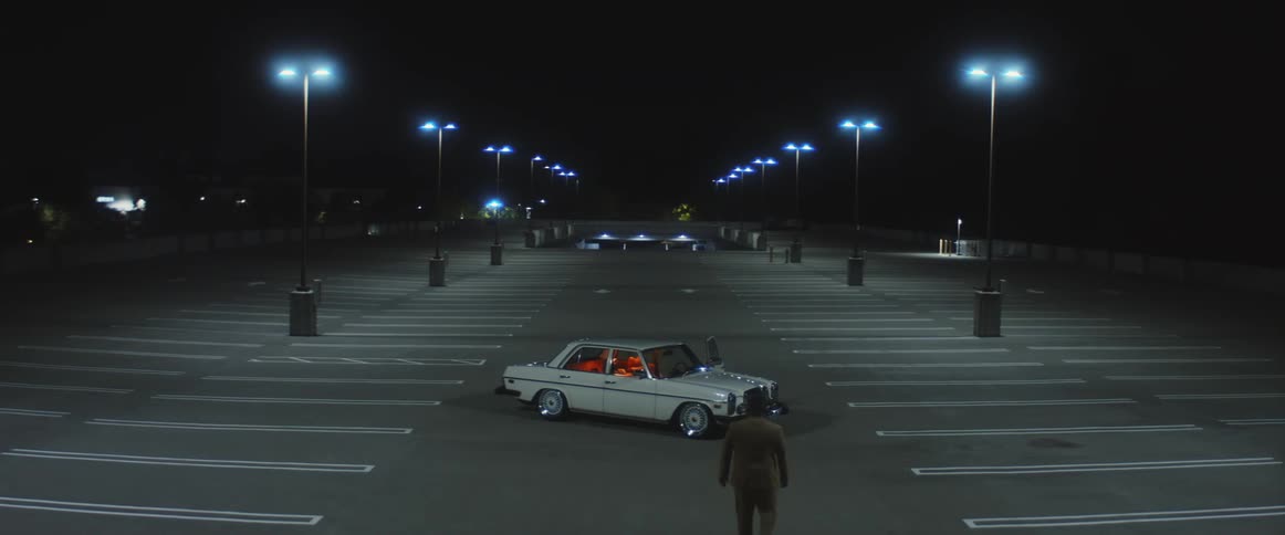 A white car parked in an empty parking lot while a man walks up to it and looks around