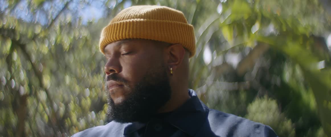 The sun shines on the face of a man wearing a coat and a yellow knit cap as his eyes are closed and then he looks up