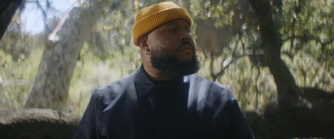 A man is wearing a coat and a yellow knit cap is standing near trees in the woods looking around