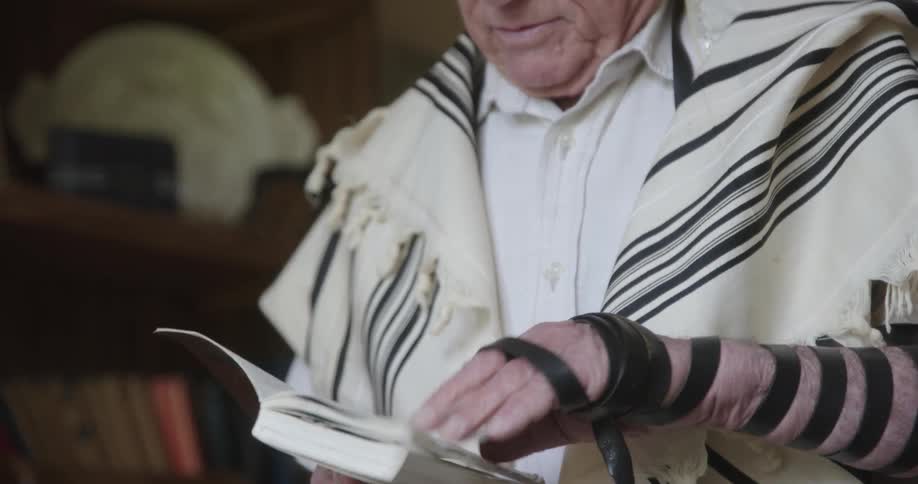 Man wearing a tefillin reads from religious book while standing near bookcase