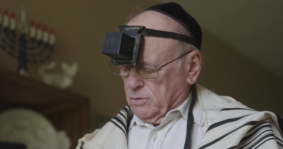Man wearing a skullcap and a tefillin reads while standing near bookcase holding a menorah