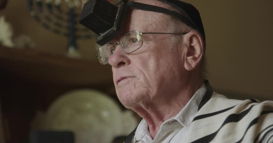 Older jewish man wearing yarmulke and prayer shawl doing morning prayers at home during the day