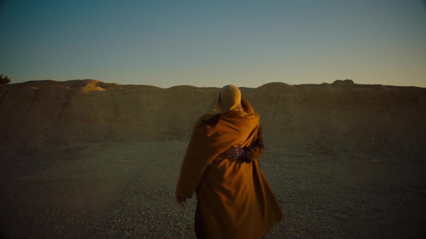 A man standing in a desert area while another man is laying down stands up and runs over to the man standing as they dance and hug