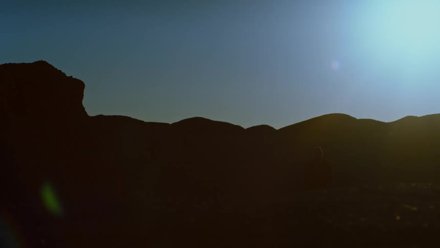 Light glares down as one person crosses the path of another person that collapses near silhouetted hills