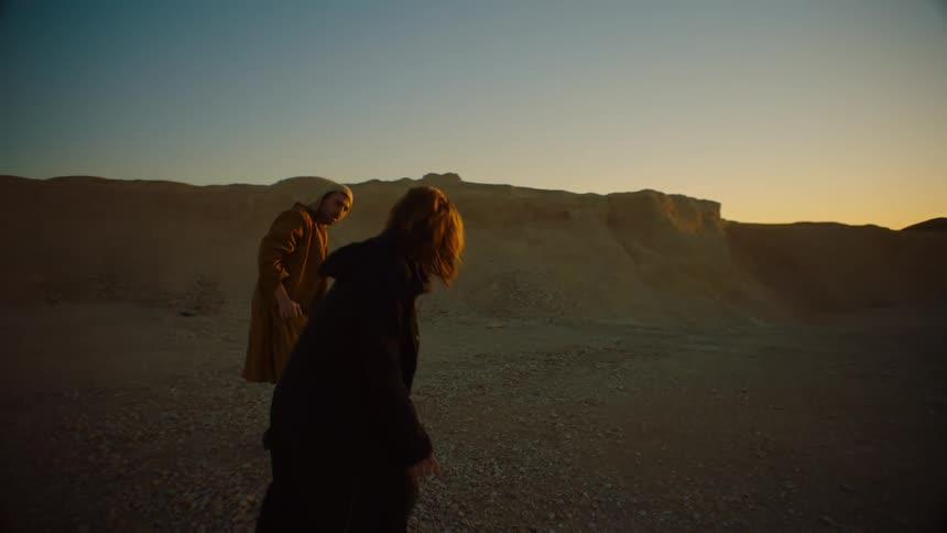 Two people slowly stand mirroring each others movements in arid plain near low cliffs under a red sky