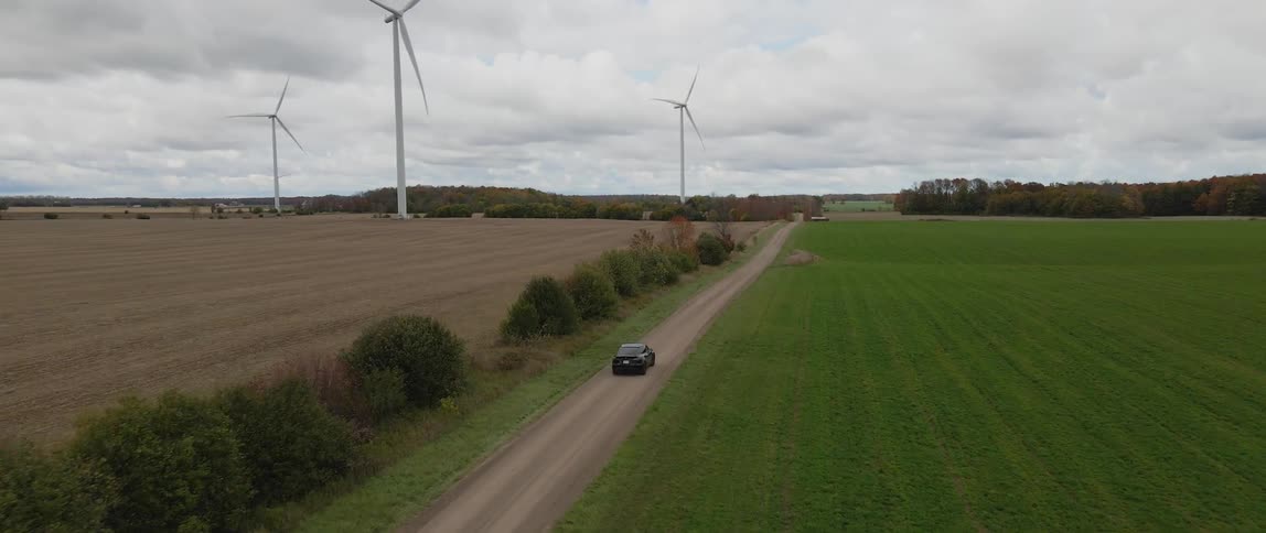 Turbines spin on a wind farm as an electric vehicle drives by on a rural road under a cloudy sky