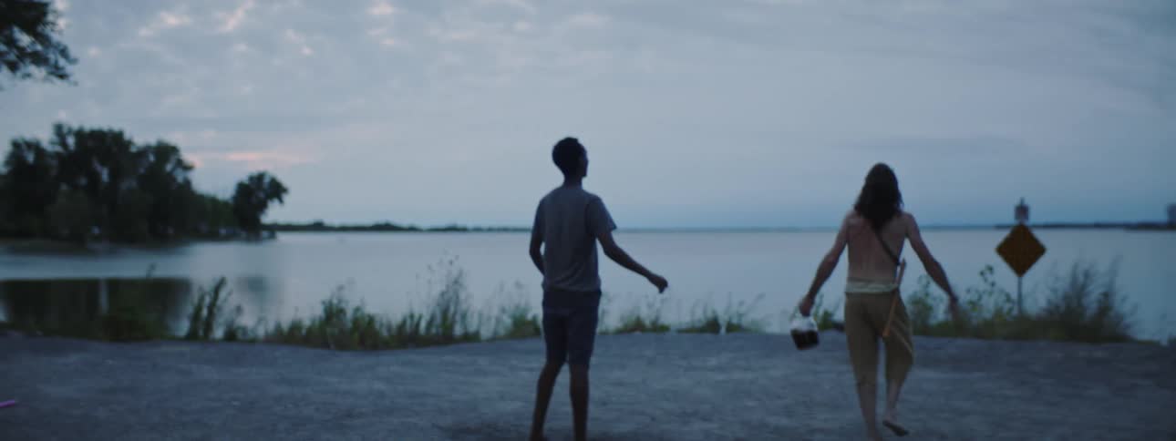Man throws hat toward water while holding bottle of alcohol as he and friend camp