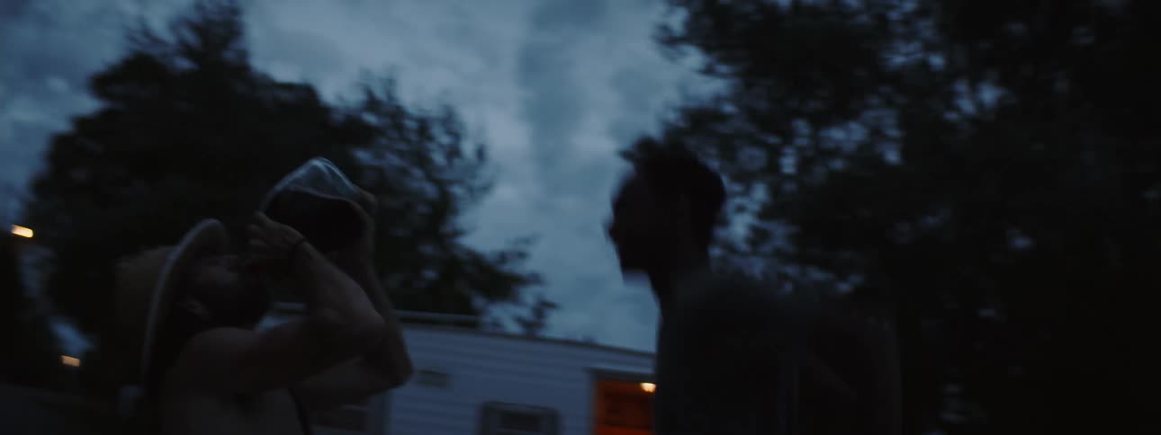 Man jumps as second man drinks from bottle of alcohol on shore with open camper in the background