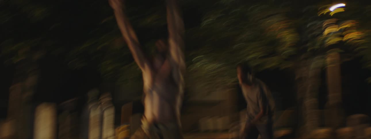 One man walking with raised hands is followed by another in a cemetery at night