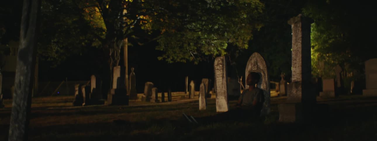 Man leans against gravestone in cemetery at night as light turns off