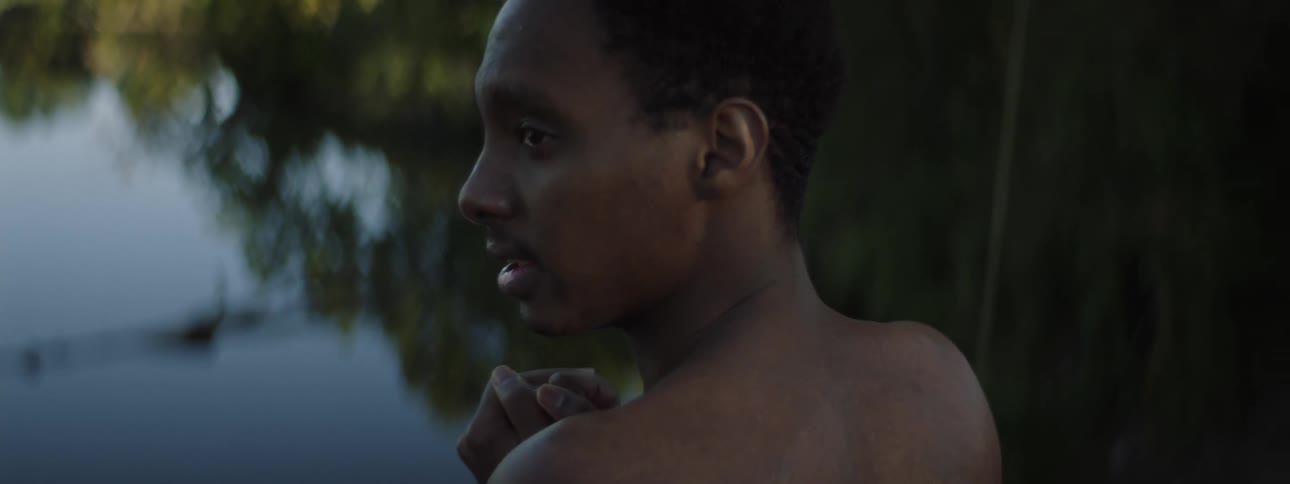The backside of a man without his shirt on stand in front of a lake as if to jump in