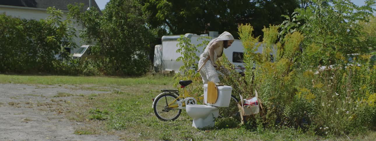 A man walking walking on a road while his friend drives his bicycle into the weeds off the road near a toilet