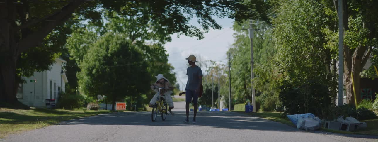 Man wearing hood with ears rides bicycle past man carrying a guitar along street