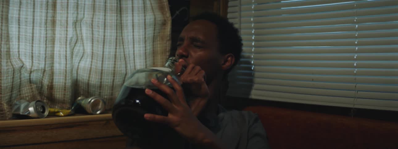 A young man sitting in the corner of a darkened room takes a swig of alcohol from a glass jug
