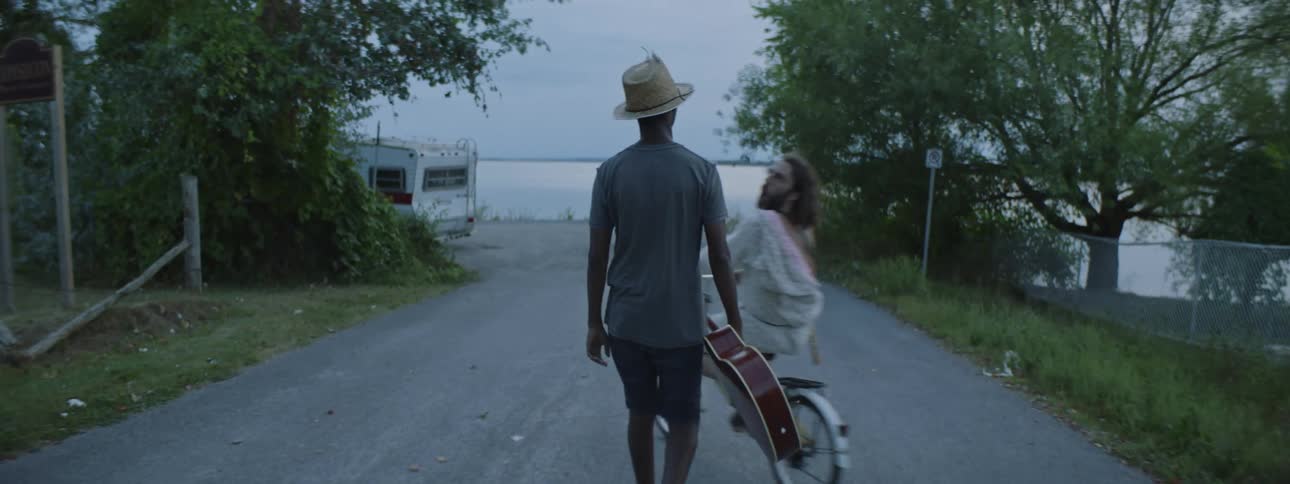 A man is walking towards the beach while holding a guitar in the background a lady rides by on her bicycle creating a dynamic moment