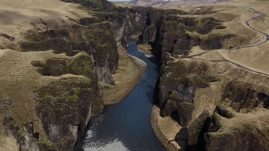 An aerial of a river in a deep canyon between cliffs as people walk a trail on top of the cliff on a plateau
