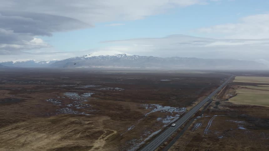 A single car travels along a roadway through a wide open landscape of fields