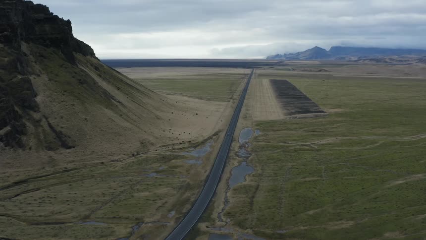 An aerial view of a car traveling on a long road through flat land surrounded by mountains on a cloudy day