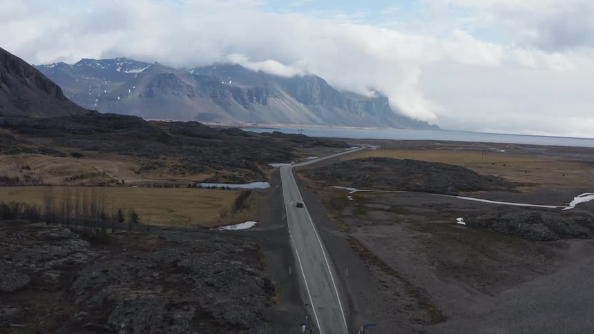 An automobile travels on a road toward the beach with mountains in the distance under a cloudy sky