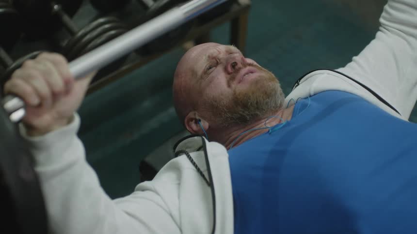 A bald man grips a weight bar while positioned on a weight bench