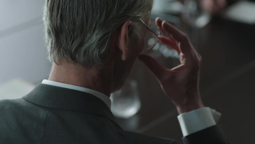 An older man wearing a suit removes his glasses while writing with a pencil