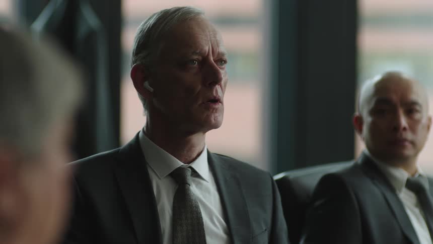 A businessman sitting next to another businessman he puts in an ear pod and turns around in his chair