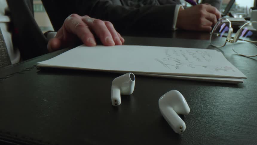 A man is writing on a notepad at a desk and then picks up an ear pod
