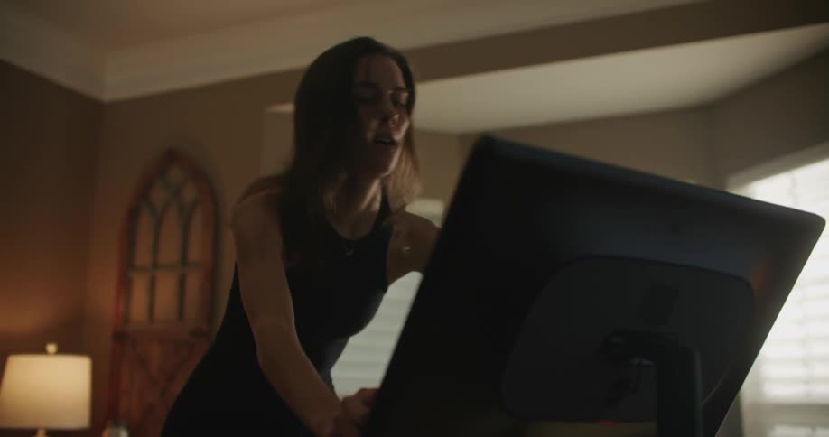 Man with brown hair black top and leggings rides stationary bike in the living room at home