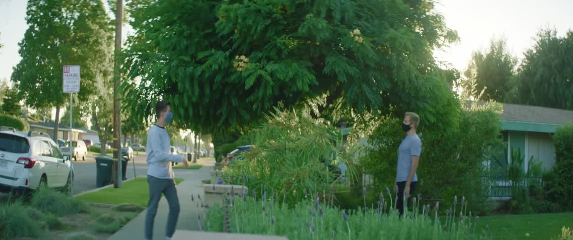 A teenager walks down a sidewalk as his friend walks down from his house and they wave and talk