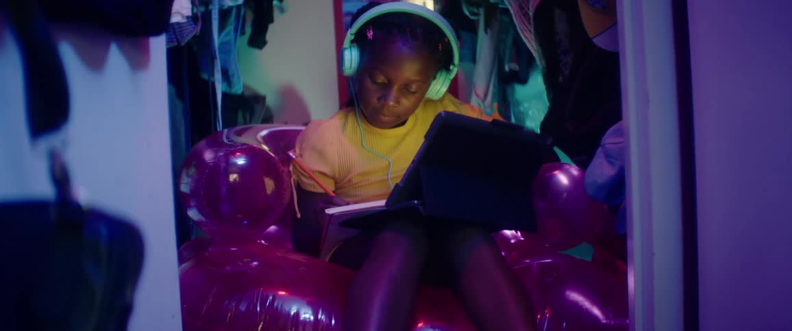 A girl sitting on an inflated couch with a pencil writing on a book in front of a laptop