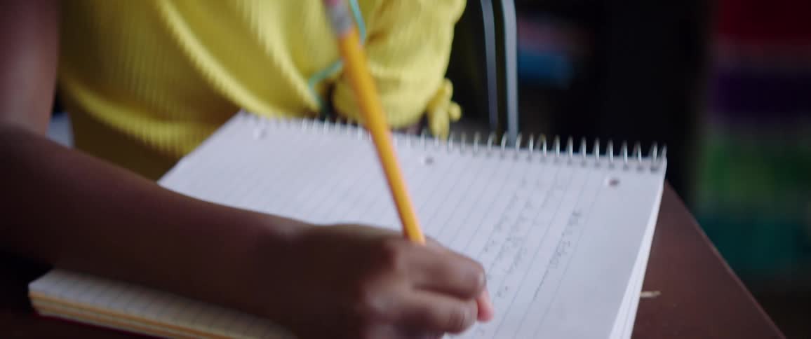 Black child with yellow shirt and turquoise headphones sitting at a desk writes with a pencil in a note book