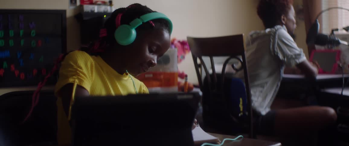 A child is sitting down writing as a woman talks into a microphone