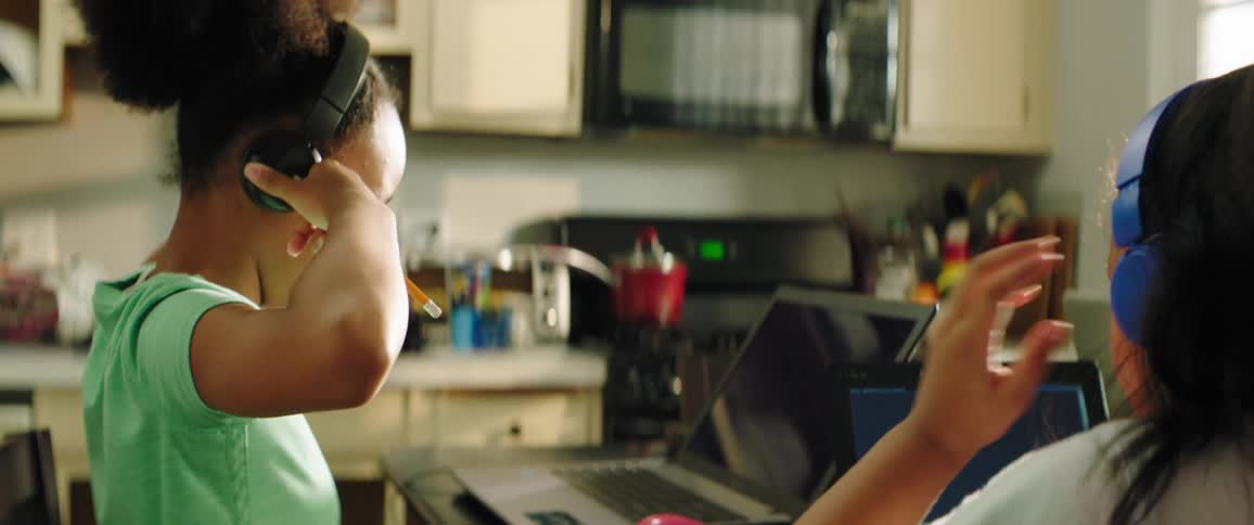 Two young girls sit at a kitchen table wearing headphones with laptops in front of them