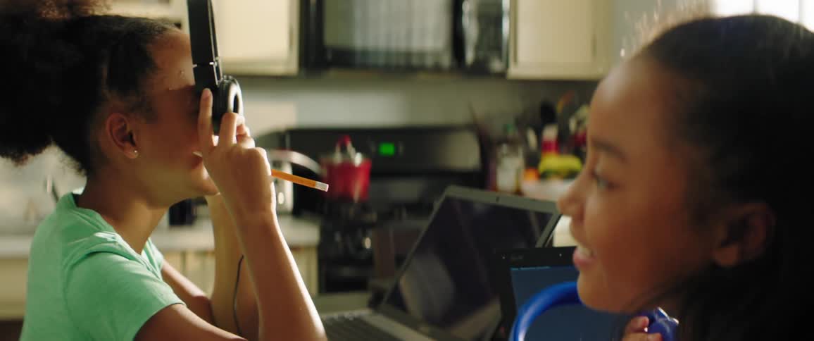 Two young females play with their headphones as they are seated in front of laptops in their kitchen at home