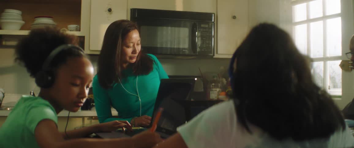 A man sits at a table with his two daughters who work on their computers and then a woman joins them