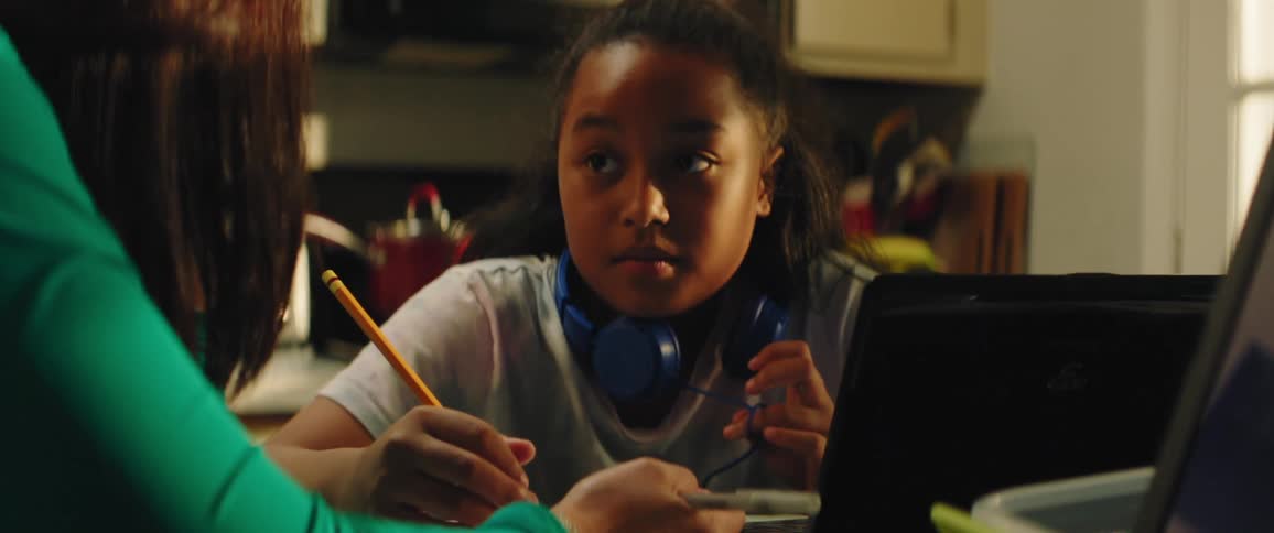 A young girl sits in front of a computer as a woman writes in a notebook and talks with the child