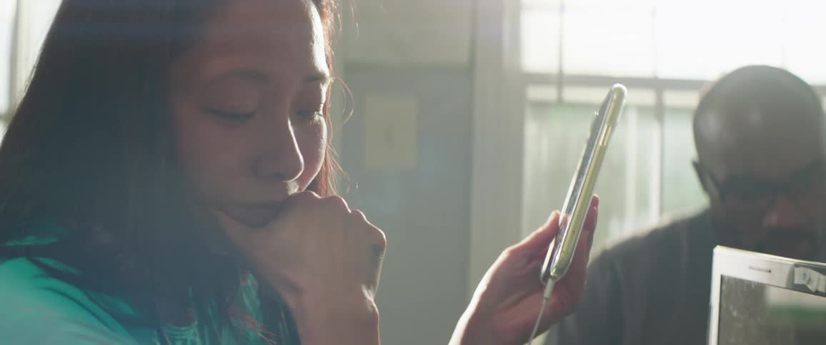 Asian woman with a turquoise shirt sitting at desk holds a smartphone and feels upset in the office