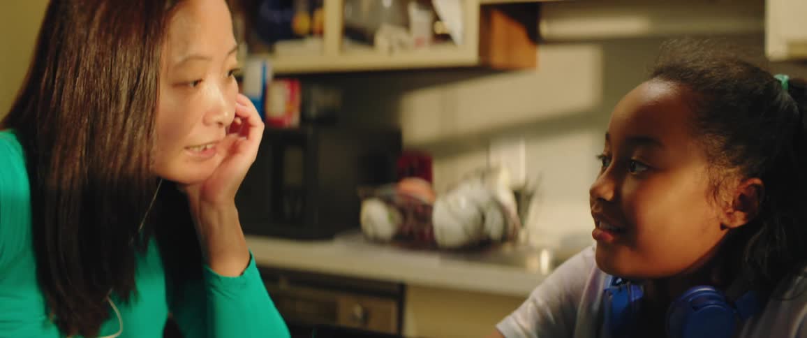 A woman and girl sit at a kitchen table with a laptop talking and laughing