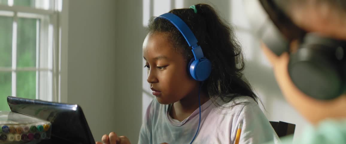 Black child wearing a white t shirt blue headphones and white child with black headphones a turquoise t shirt sitting at a table uses a laptop and writes in a notebook in a room during daylight