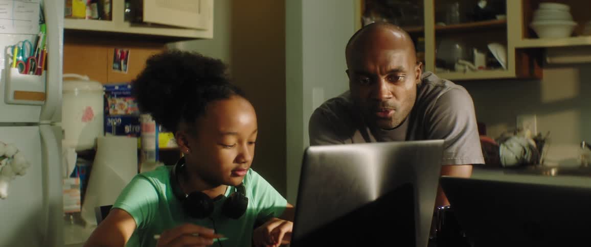 Daughter with a turquoise t shirt sits at the table and uses a laptop while father with grey t shirt drinks a cup coffee and checks her homework