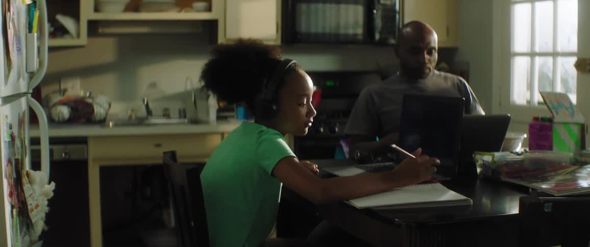 Child with headphones and a turquoise t shirt sitting at the table uses a laptop and writes down while father with a grey t shirt uses a laptop and drinks a cup of coffee in the kitchen at home
