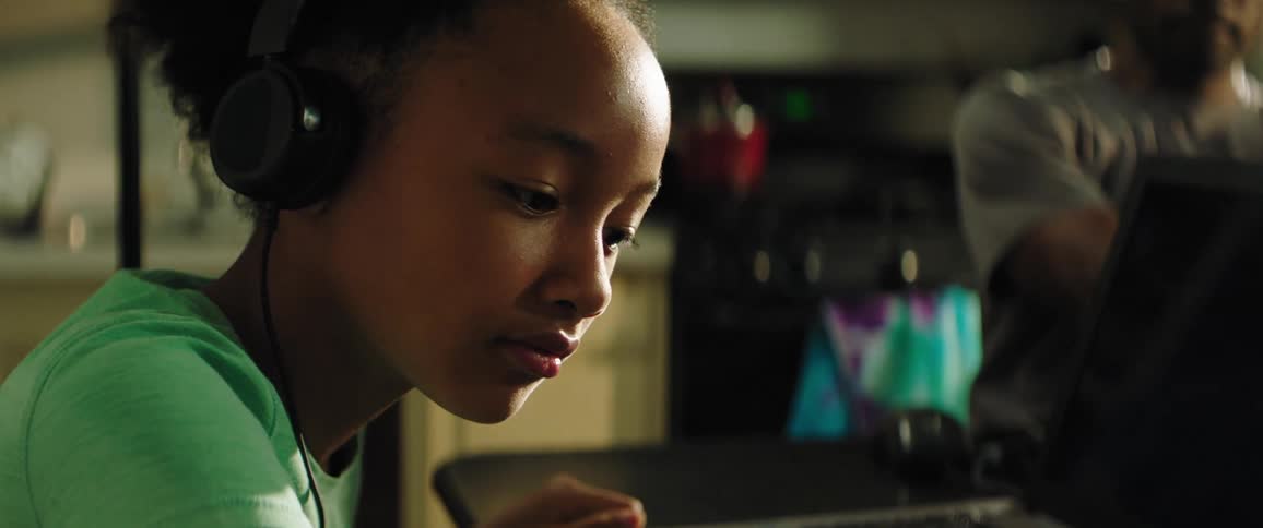 Daughter with a turquoise t shirt and black headphones sits at the table does homework while father with grey t shirt drinks a cup coffee and uses a laptop in the kitchen at home