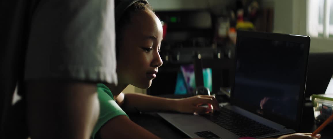 A girl works on a laptop as a man eats in the kitchen