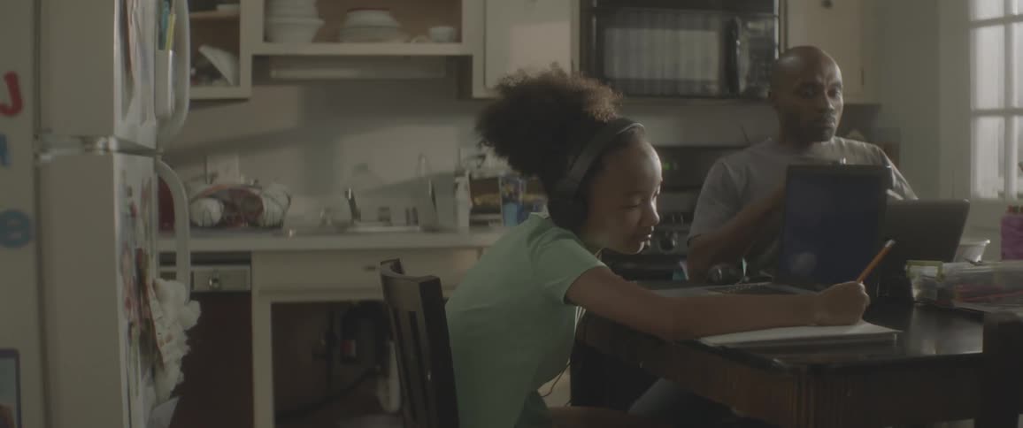 Child with headphones and a turquoise t shirt sitting at the table uses a laptop and writes down while father with a grey t shirt uses a laptop and drinks a cup of coffee in the kitchen at home