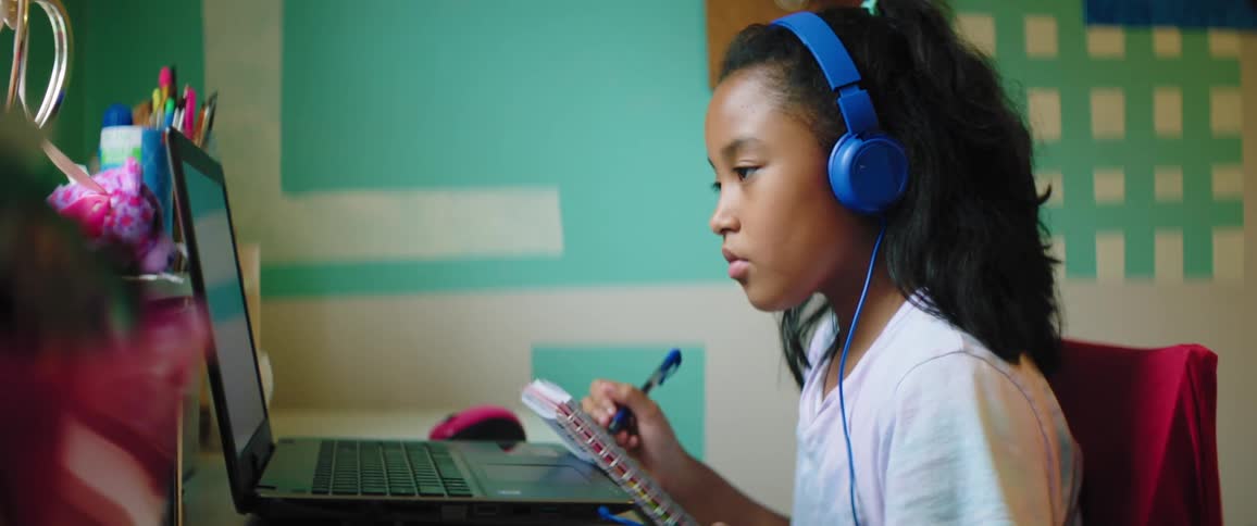Black child wearing blue headphones sitting at a desk looks at a laptop and takes notes in a room