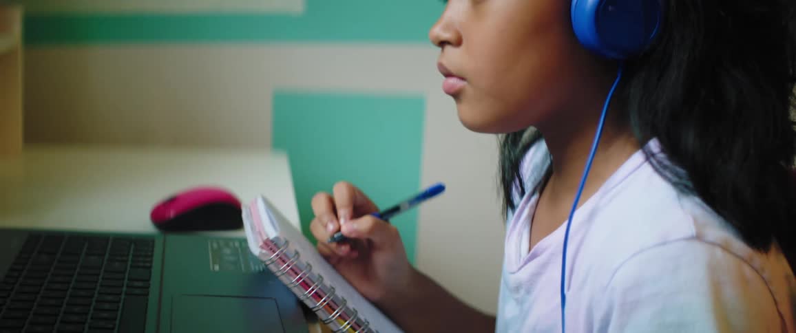 Child with a t shirt and blue headphones looks at a laptop and writes in a notebook