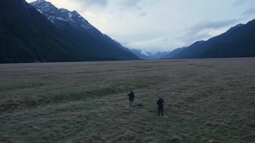 Two people are standing near tripods surrounded by mountains on a field