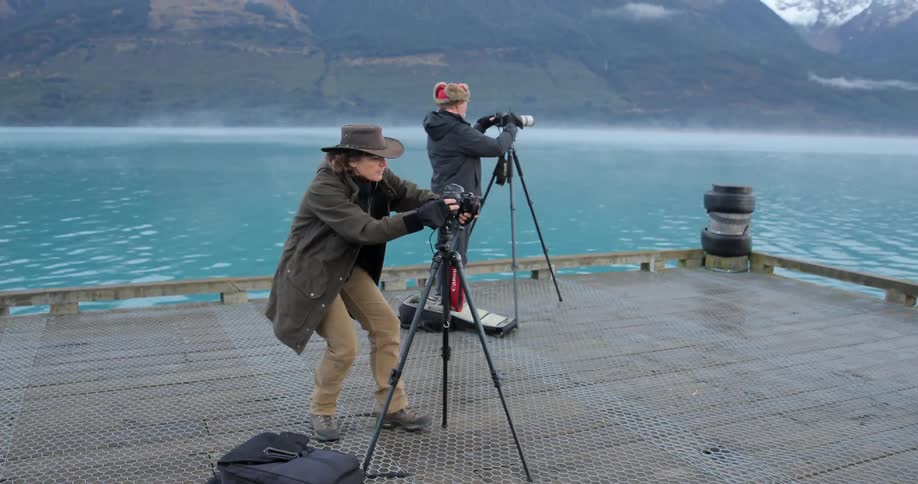 Two people are using cameras on a pier near a mountain