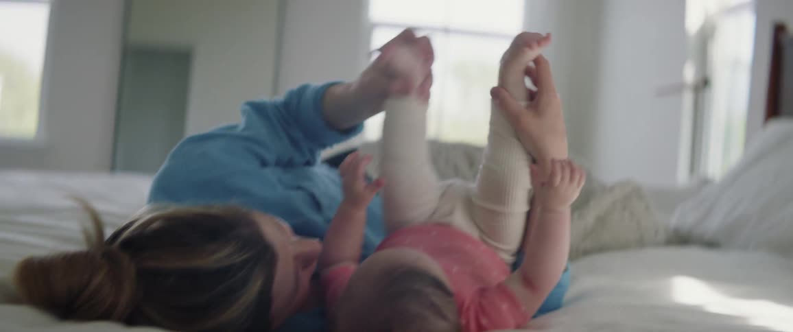 Mother and infant daughter laying on a bed playing as the mother holds the babies legs