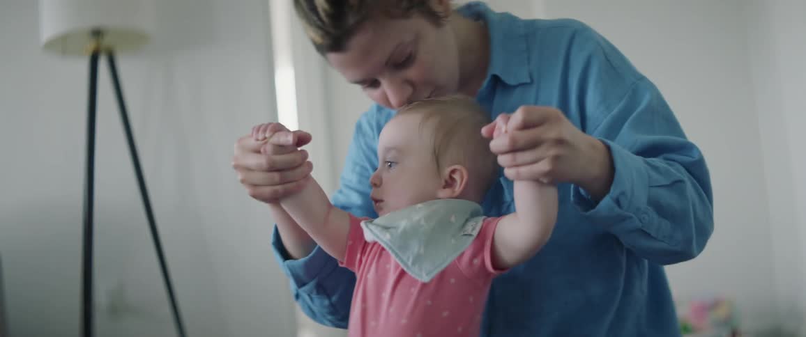 A woman is kneeling on the floor as she holds her babys hands in the air teaching her how to stand and walk